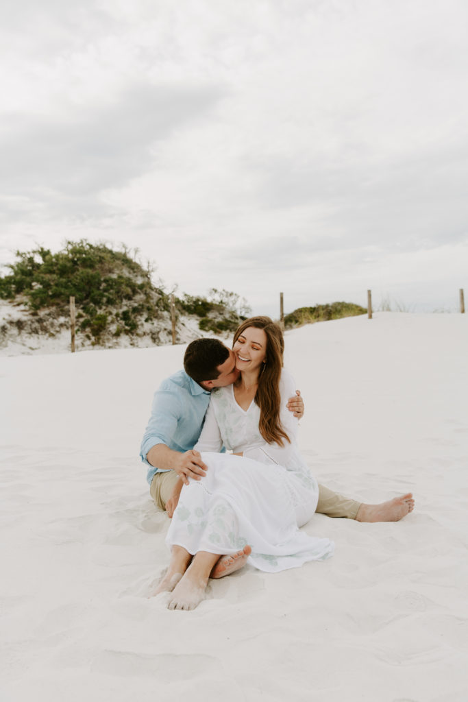 A couple sitting in the sand with the woman in her partners lap as he blows raspberries into her neck making her laugh during their beach engagement photos in Florida