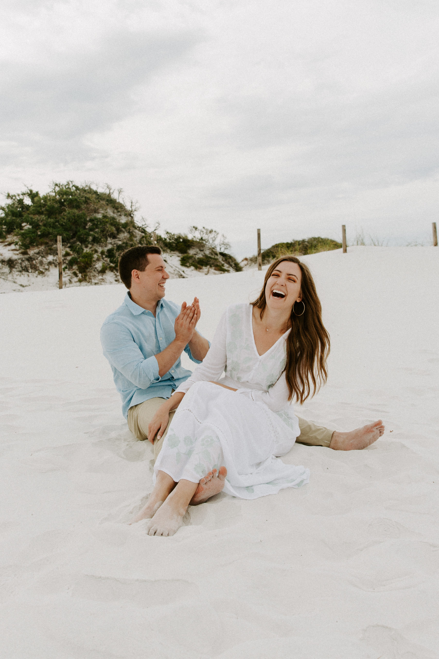 Couple sitting in the sand laughing as the man is clasping his hands together at something his partner said during their Florida engagement photos