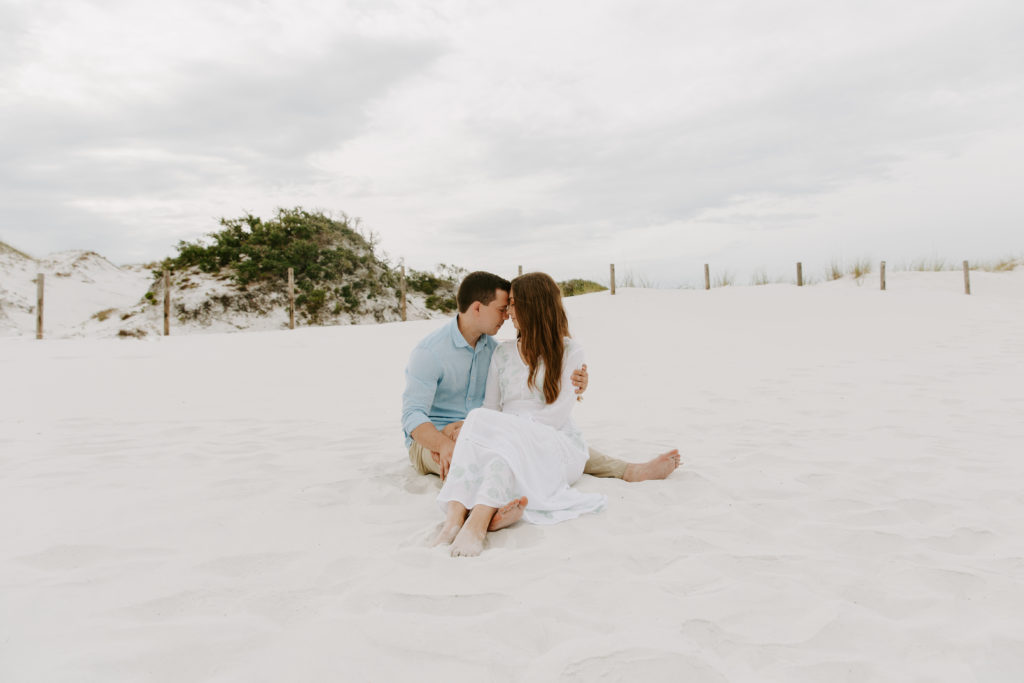 A couple sitting in the sand with the woman in her partners lap cuddling up to him with their foreheads together during their Florida panhandle engagement photos