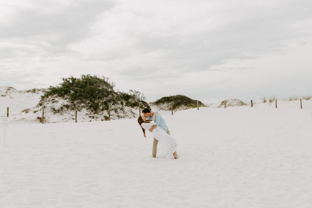 A man dipping his partner to give her a kiss with the large sand dunes behind them during their Florida beach photos