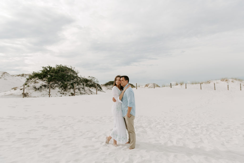 A couple holding onto each other as they both look off into the distance as the sun rises and the sand dunes rise behind them during their emerald coast engagement photos
