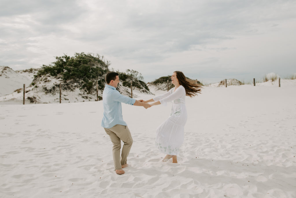 A couple holding hands and spinning in circles as the woman's hair is flying out behind her and they are both laughing with sand dunes in the background during their Florida panhandle engagement photos