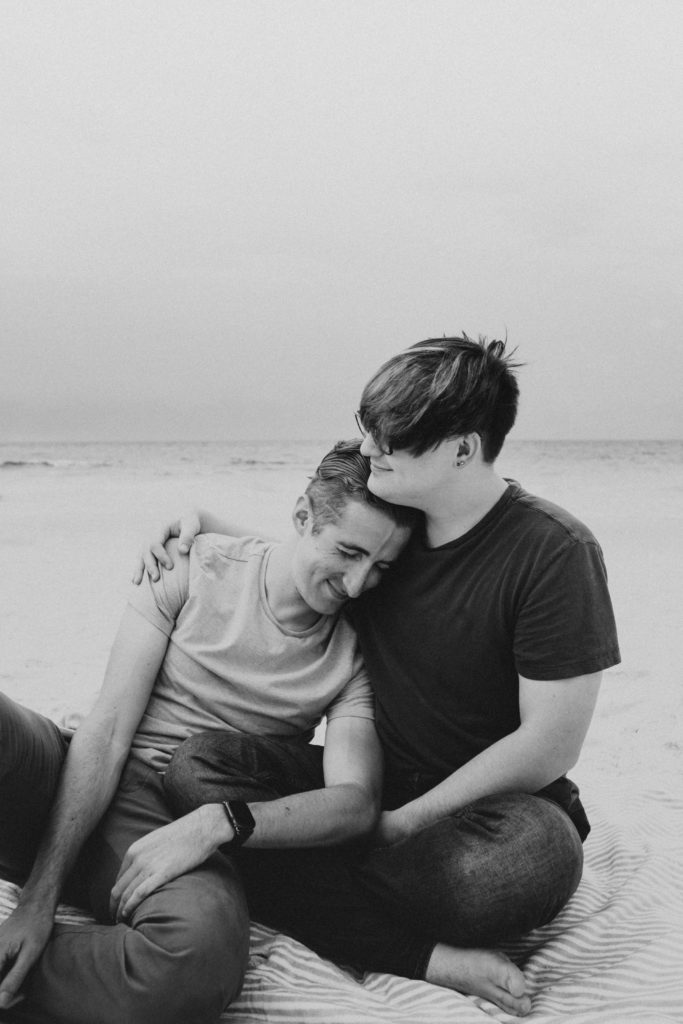 A couple sitting down on the beach as one partner leans into his partner with their arms around each other