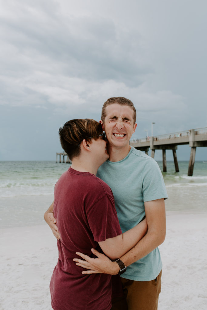 Man blowing kisses into his partners neck as they both laugh with a pier in the background