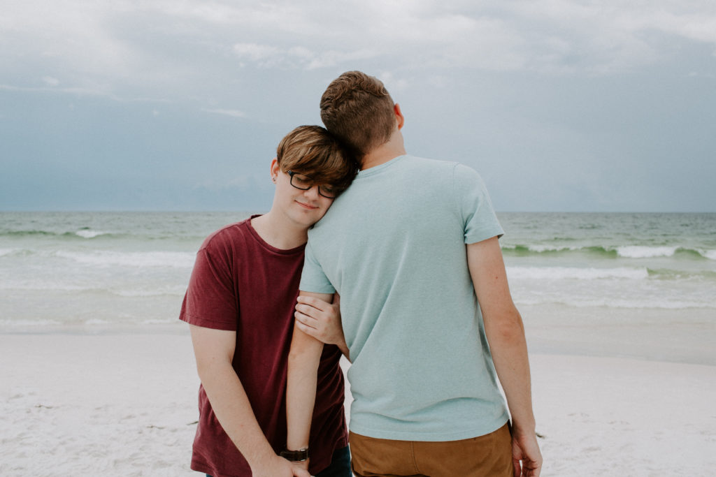 Couple standing on the beach with the first partner facing the ocean and the second one facing away holding his hand