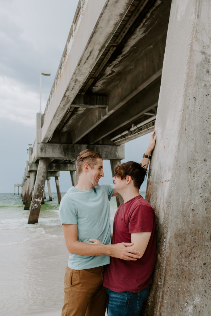 Couple leaning up against the base of the pier looking at each other and smiling during their early morning Okaloosa Island beach session