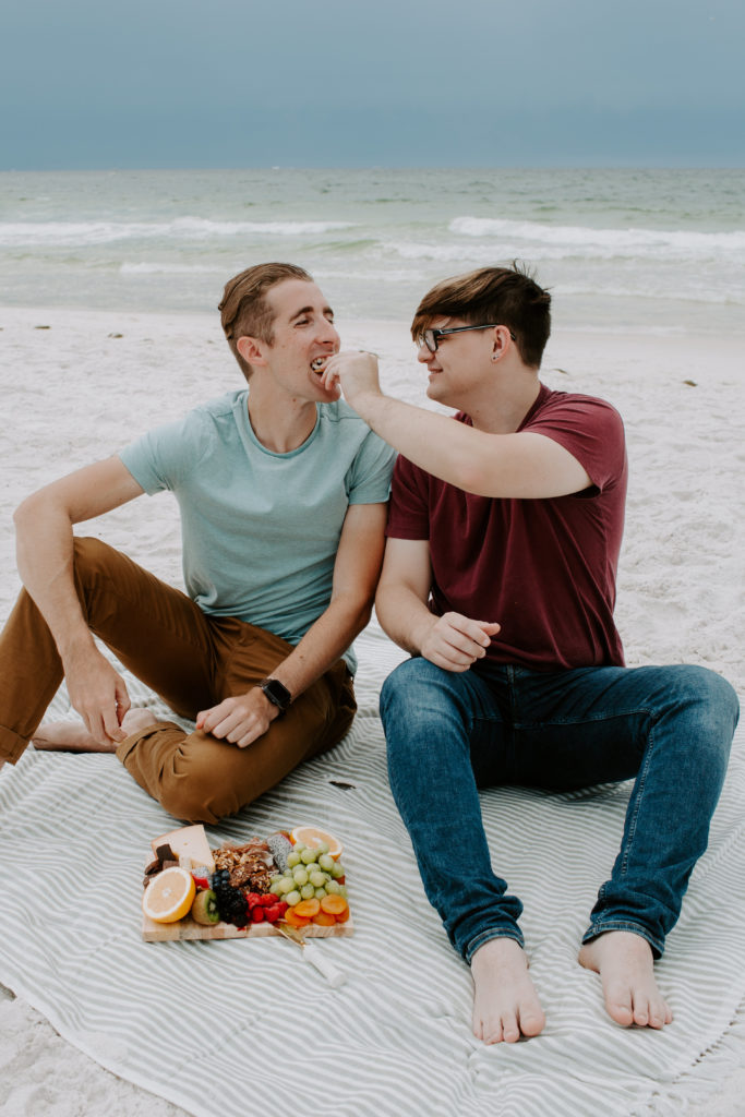A couple sitting on a beach blanket with a charcuterie board while one partner feeds the other partner grapes during their morning photoshoot