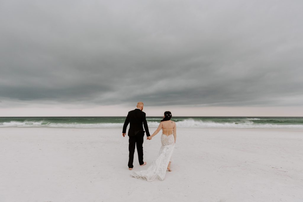 Couple holding hands and walking towards the ocean that is getting darker as the storm comes into