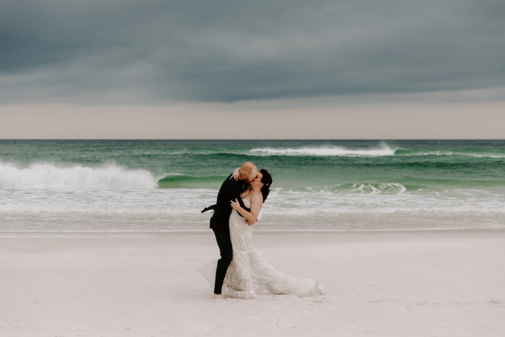 Couple sharing a kiss with wind blowing his jacket different directions during their beach elopement