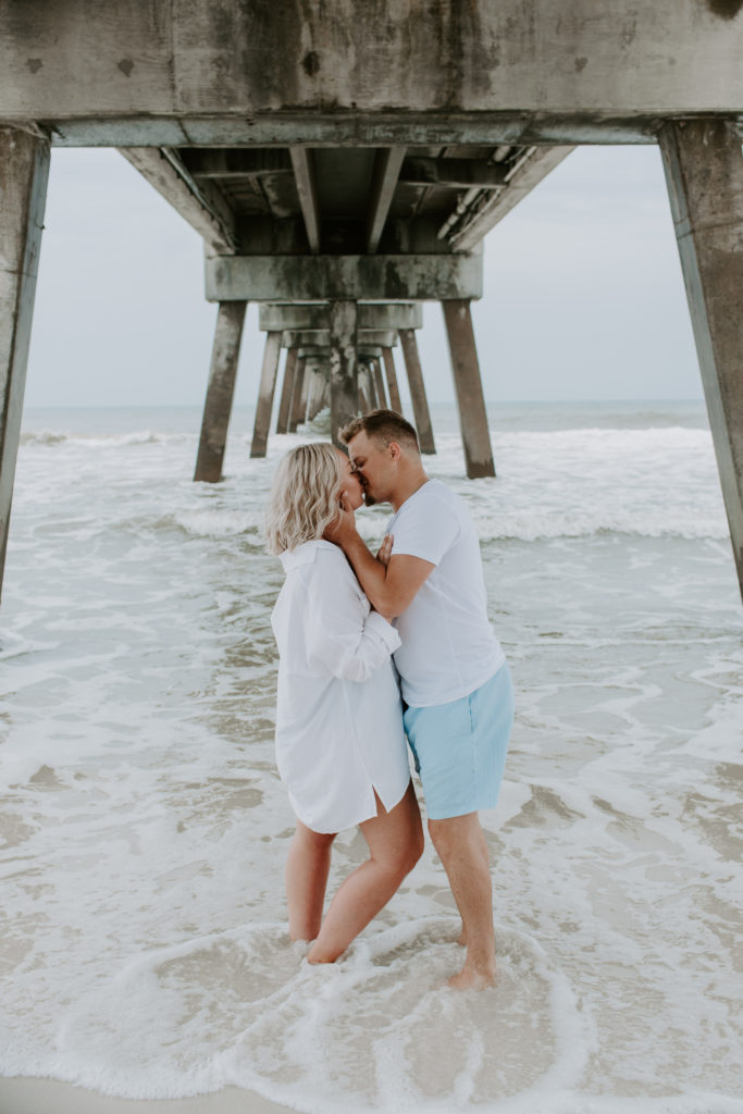 Couple standing under a pier sharing a kiss during their morning beach photoshoot