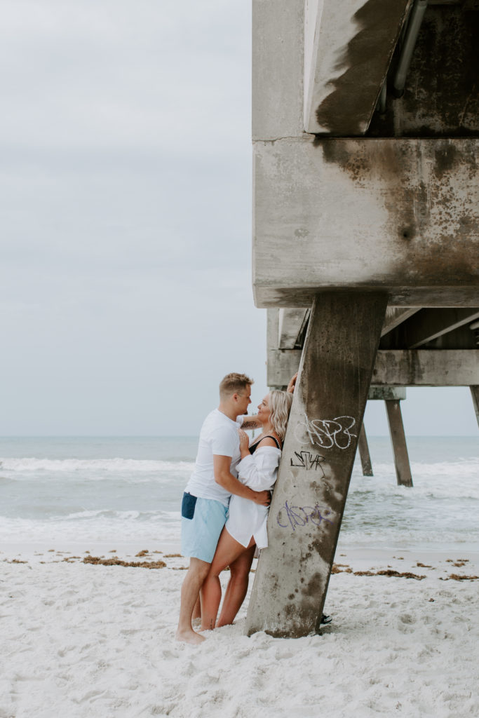 Couple leaning up against a pier looking into each others eyes with the calm ocean in the background
