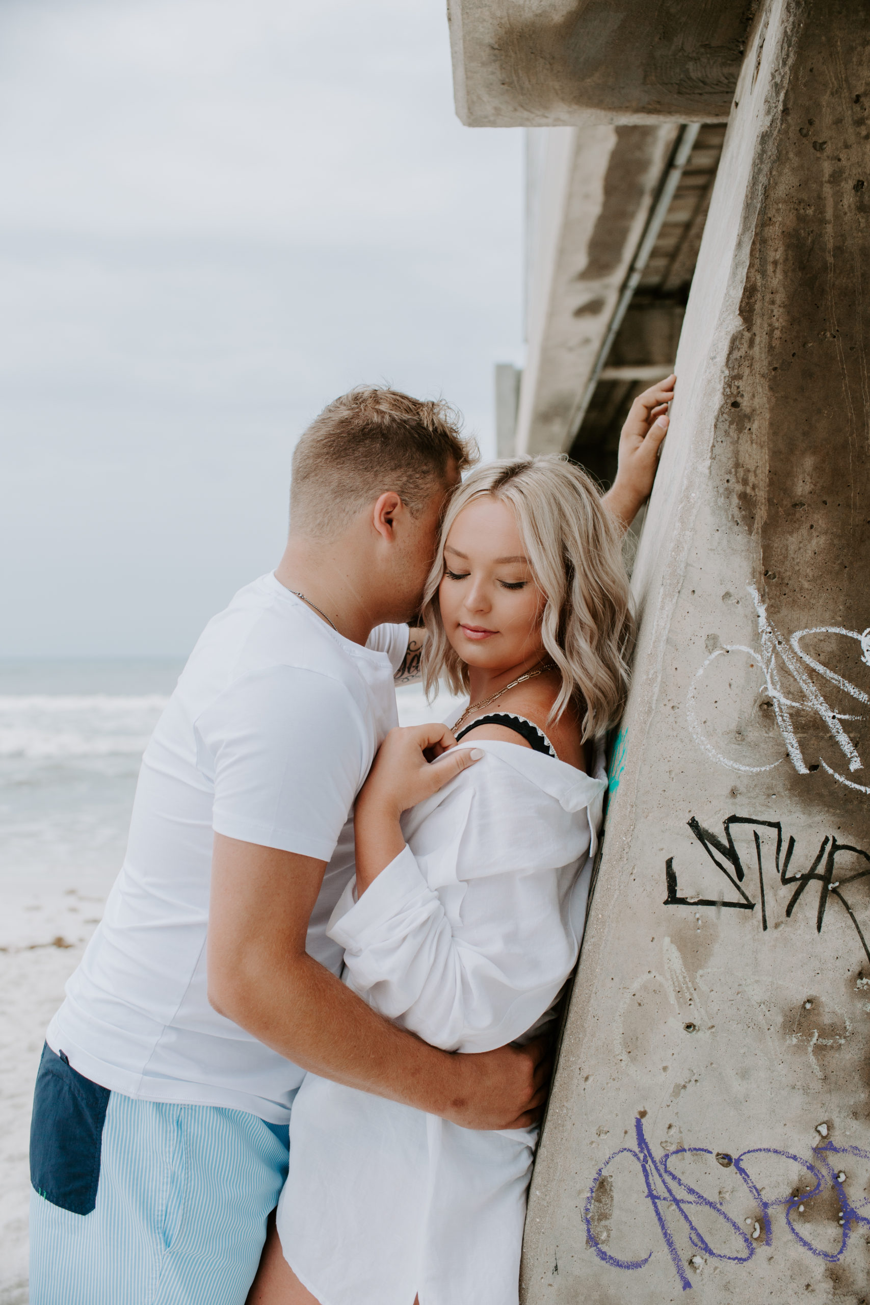 Couple leaning up against a fishing pier with the woman holding up the edge of her coverup and the man is giving kisses into the side of her head with his arm leaning against the pier during their beach engagement photos