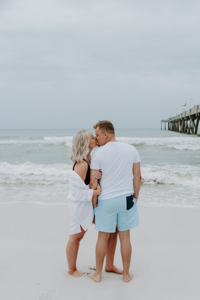 Couple standing on the beach sharing a kiss as the woman holds onto the mans arm and he is facing the ocean during their Destin, Florida engagement photos