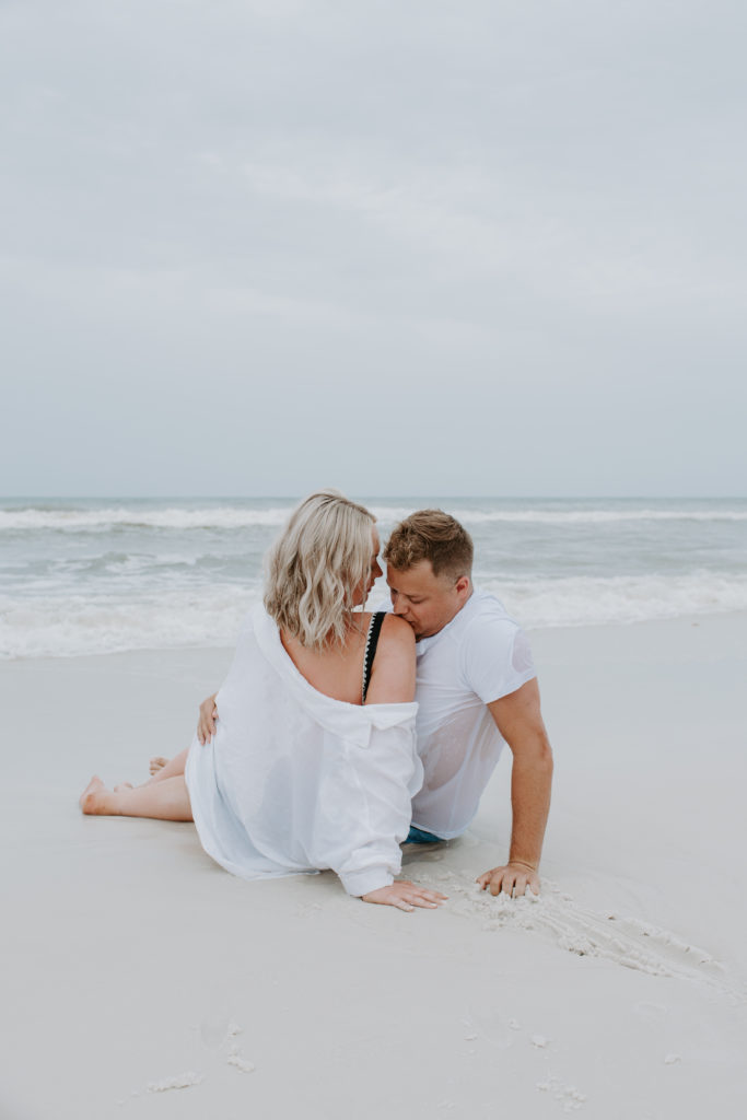 Couple sitting on the beach facing each other as the man kisses his partners shoulder during their early morning engagement session