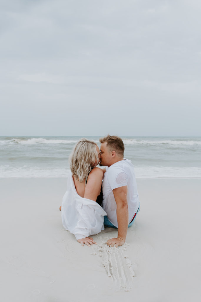 Couple sitting on the beach propping themselves up with their arms as they share a kiss during their sunrise beach photoshoot