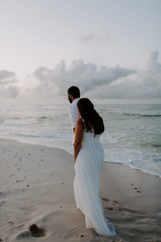 A man leading his partner along the ocean as she holds up her dress during their sunrise beach photos
