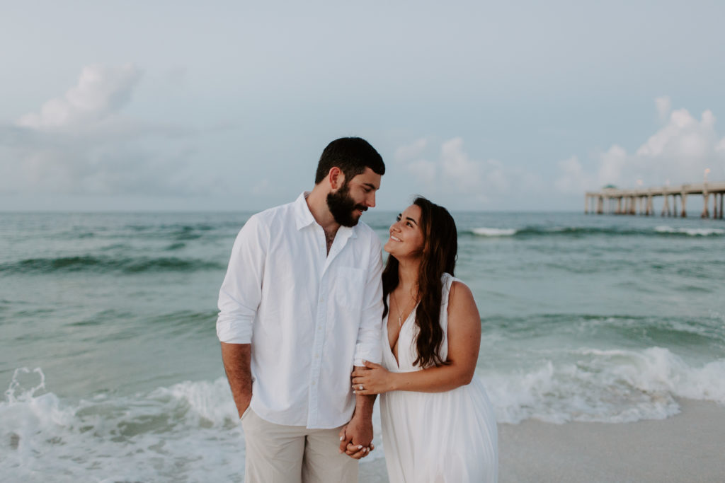 A woman holding onto her partners arm as she looks up him and he looks down at her during their sunrise beach engagement photos