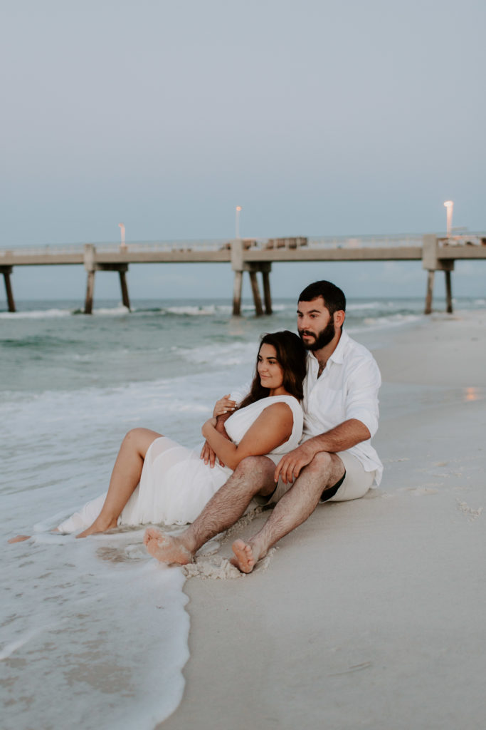 A couple sitting in the sand with the woman leaning back into the man as the man has his arms around her and they are both looking off into the distance
