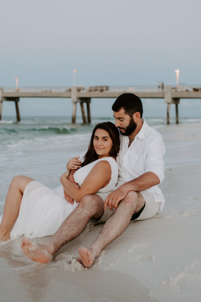 A couple sitting in the sand with the woman leaning back into the man and he is looking down at her during their beach engagement photos