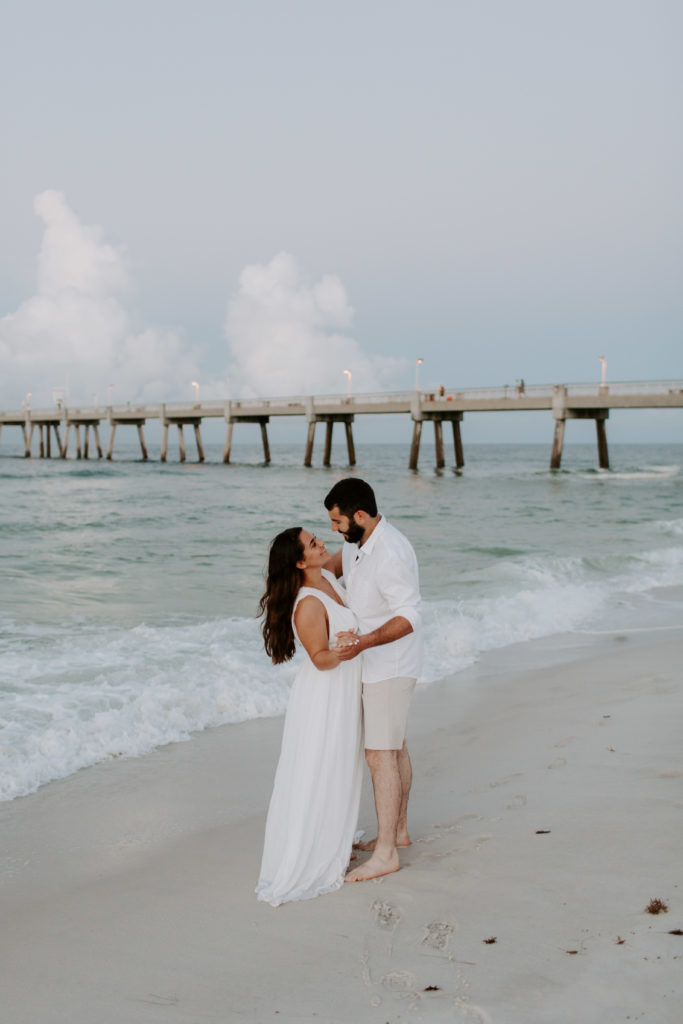A couple standing on the beach looking at each other with the Okaloosa Island Pier in the background during their sunrise engagement photos