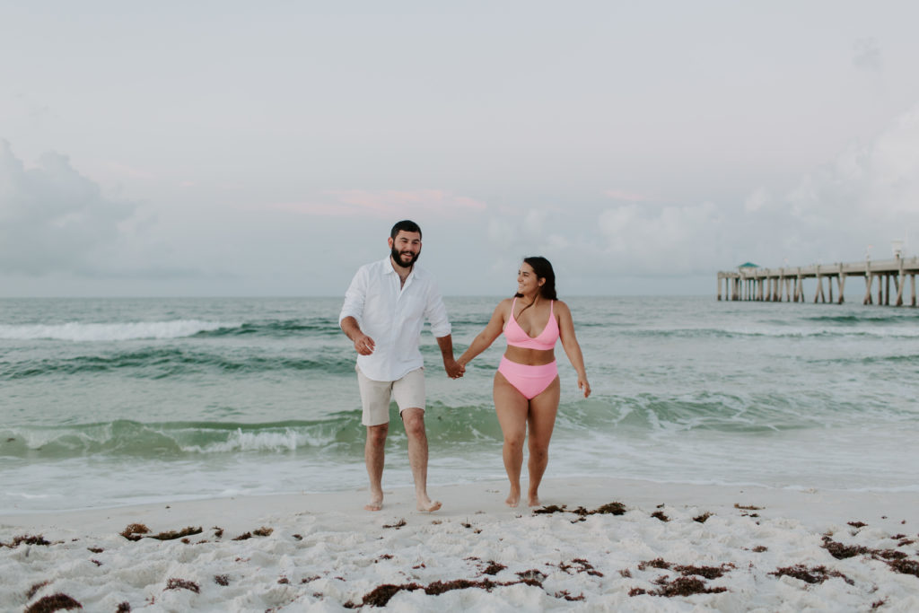 Couple holding hands and running away form the ocean as the woman looks at her partner smiling during their Destin engagement photos