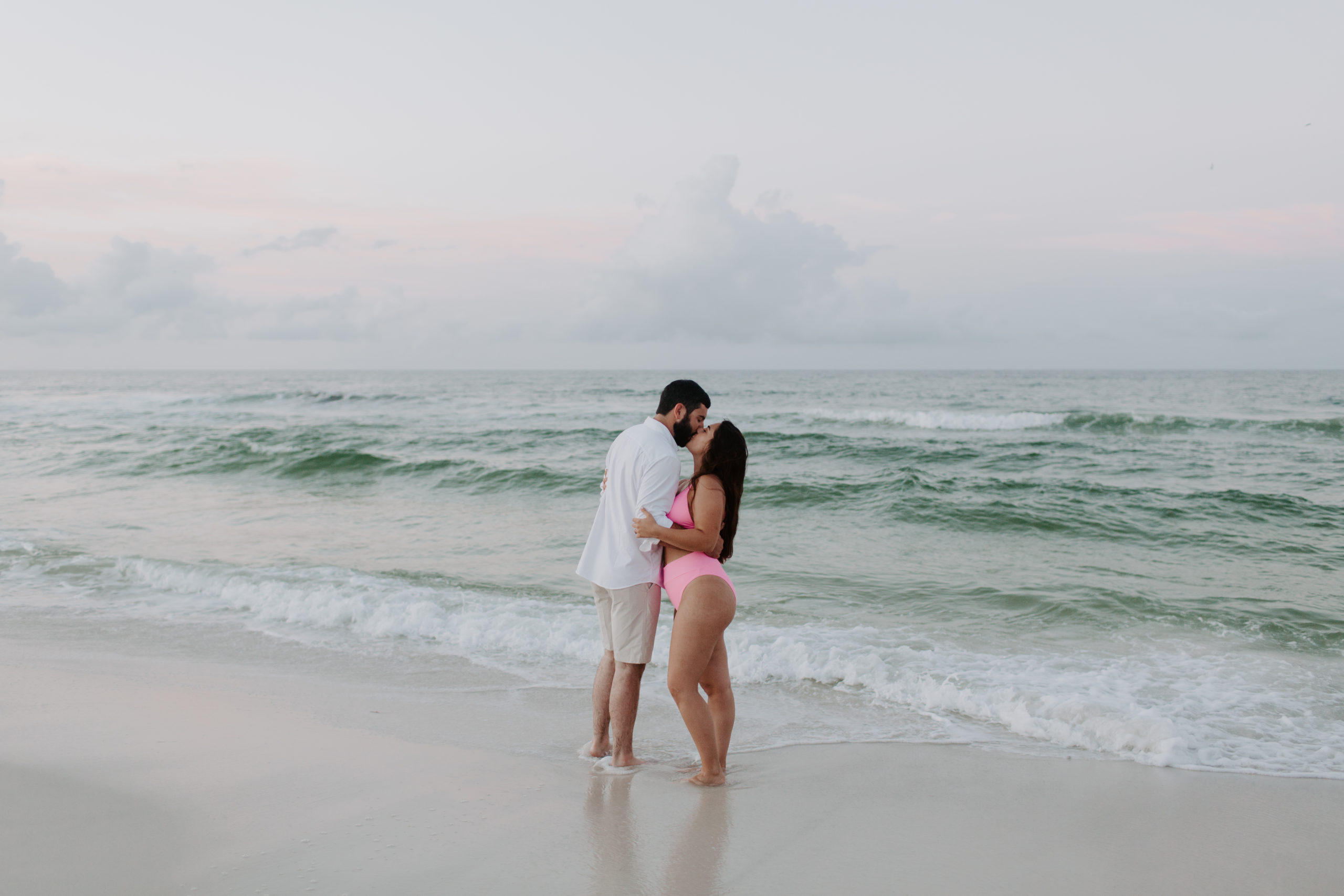 A man and a woman standing at the ocean line in their swimsuits sharing a kiss as the sun rises