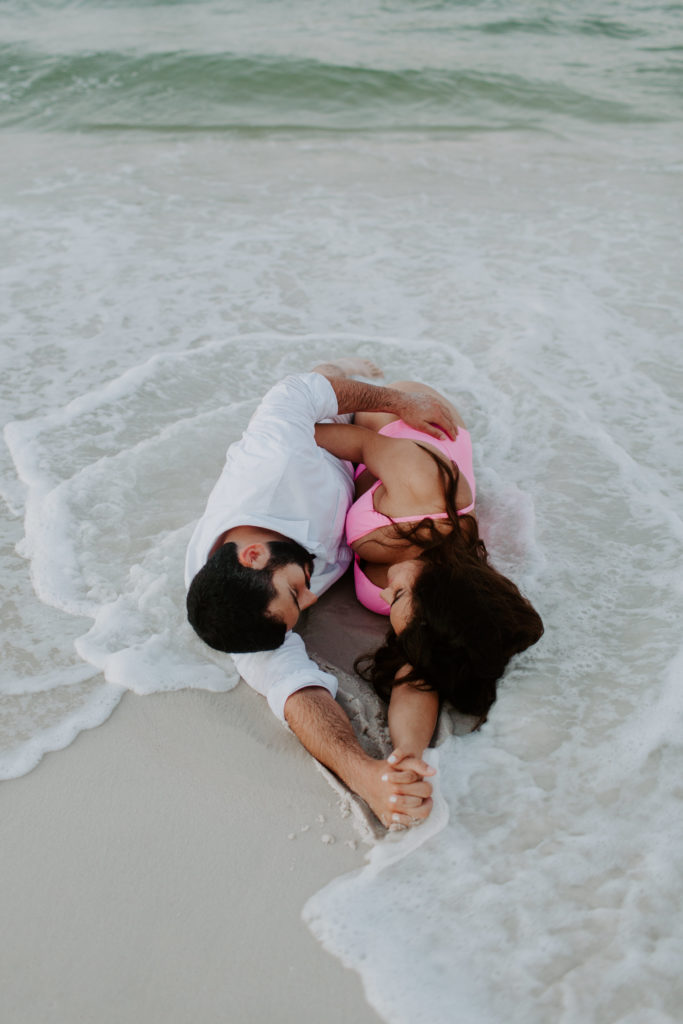 Couple laying down in the sand on the beach as the water washes up over them and they are holding hands and facing each other