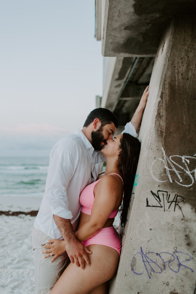 Couple leaning up against a wall as they share a kiss with the woman in a pink swimsuit during their early morning photoshoot in Destin, Florida