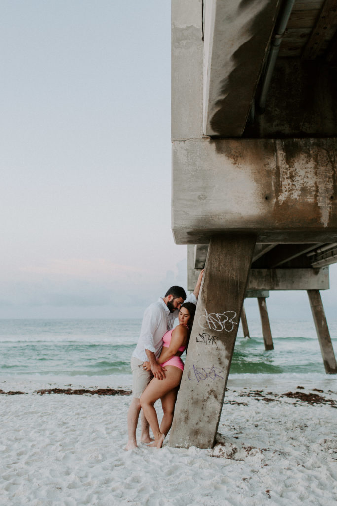 Couple leaning up against a fishing pier as the woman looks down at the sand and her partner looks down at her during their sunrise engagement photos in Florida