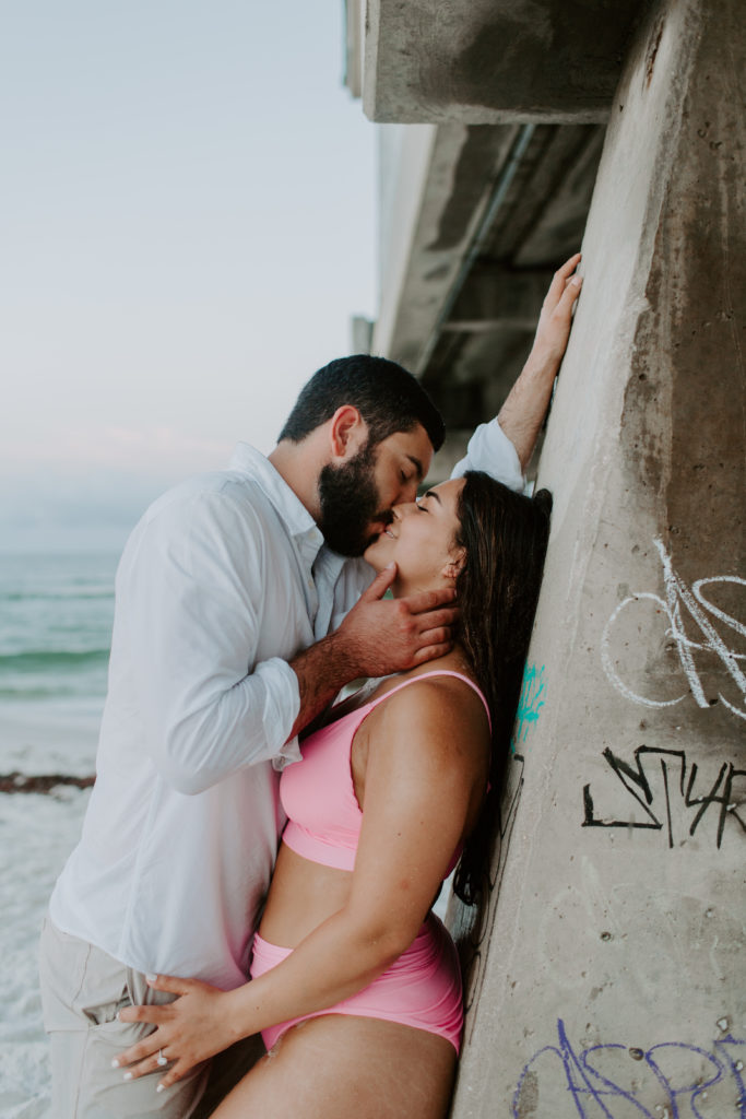 Couple leaning up against the side of a pier as the man brings his partner in for a kiss during their sunrise beach photoshoot