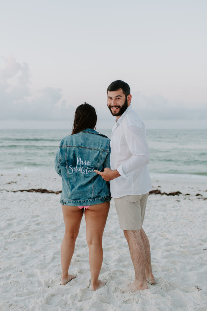 A woman facing the ocean as her future husband points at the back of her jacket during their beach sunrise couple session on Okaloosa Island