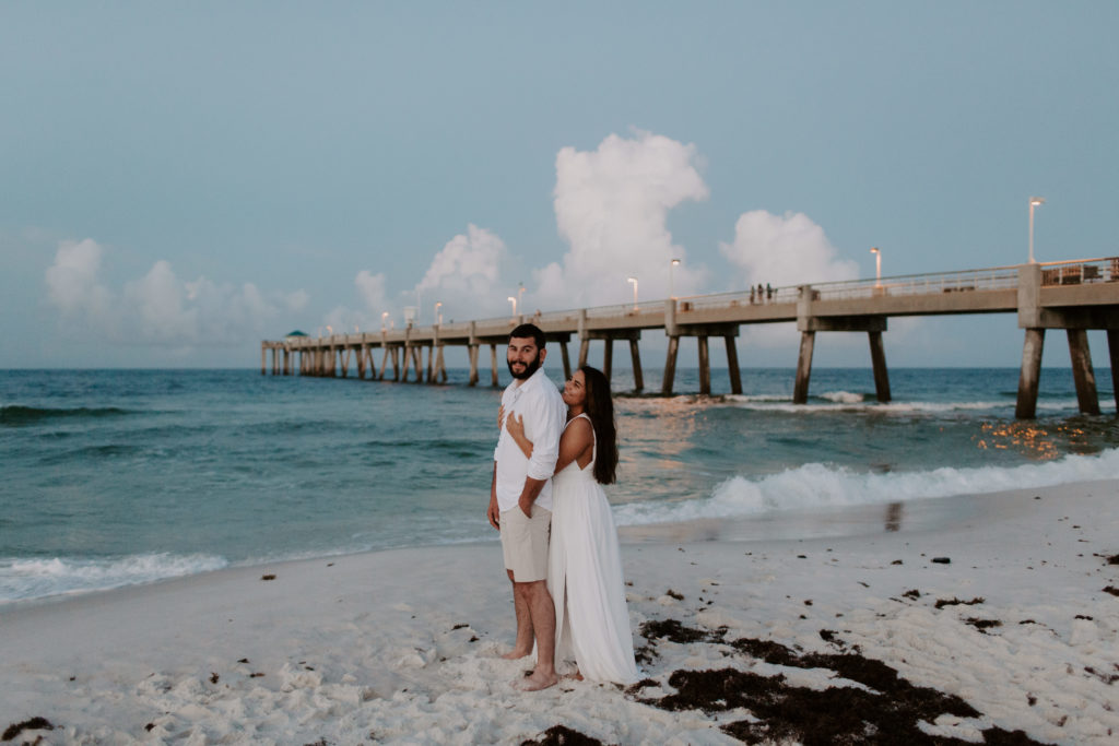 A couple standing along the water line with the woman standing behind her partner holding her arms around him and looking up at him as he looks of into the distance with the Okaloosa Island pier in the background