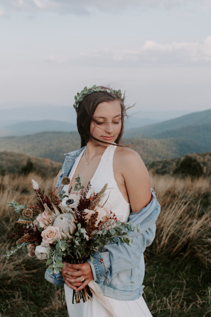 woman holding her bouquet looking down at the ground as the wind whips her hair around her face during her elopement at sunrise in the north carolina mountains