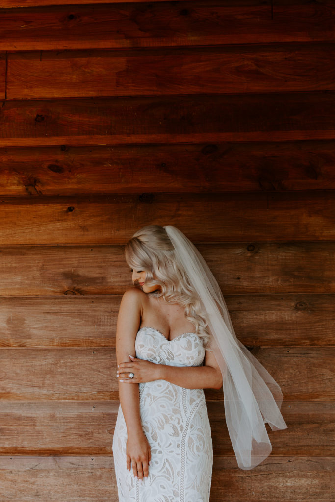 Bride holding onto her arm as she looks down at her shoulder with her veil blowing in the wind as she is standing in front of the cabin wall during their Arkansas elopement