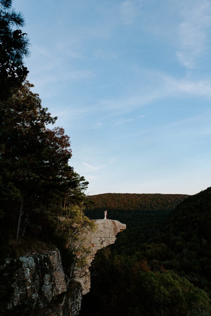 Couple standing on a cliff overlooking a valley with trees Turing different colors during their Arkansas elopement in the Ozarks