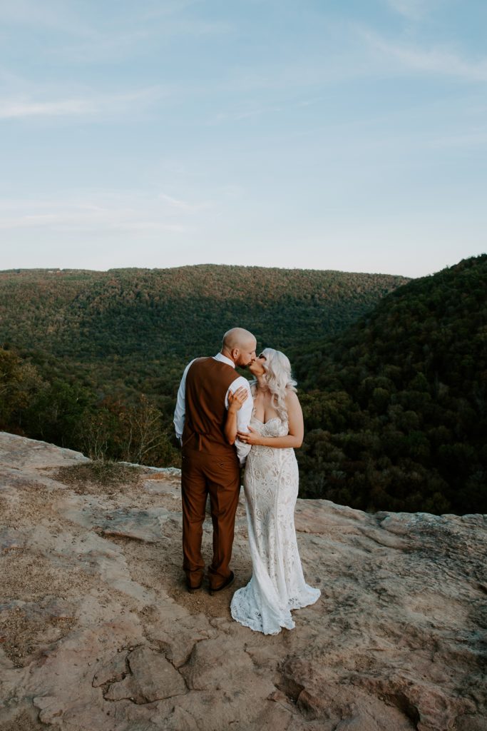 Couple standing on the edge of cliffs with the fall colors behind them sharing a kiss as the woman holds on to her partner during their hiking elopement