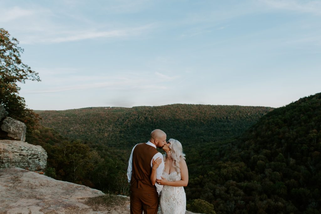 Couple standing on the edge of cliffs with the fall colors behind them sharing a kiss as the woman holds on to her partner during their Arkansas elopement