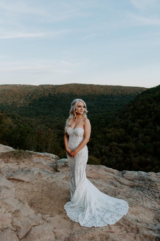 Woman standing on the edge of the cliffs holding her arms in front of her looking off into the distance in her lace wedding dress during her Ozarks elopement