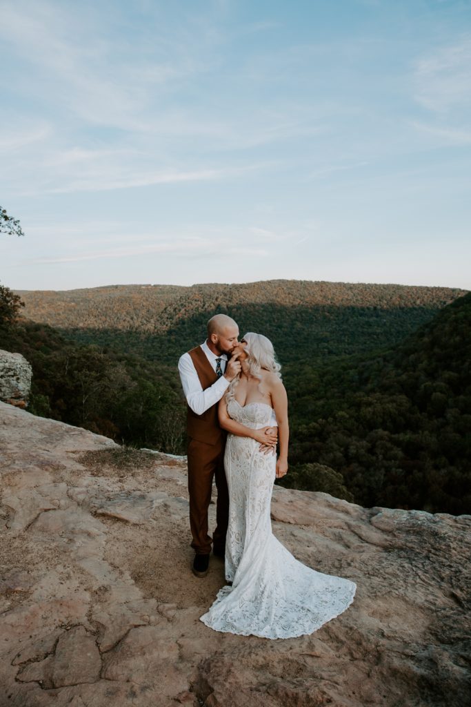 A man gently giving his partner a a kiss while they are holding hands with the Ozarks in the background during their Arkansas elopement