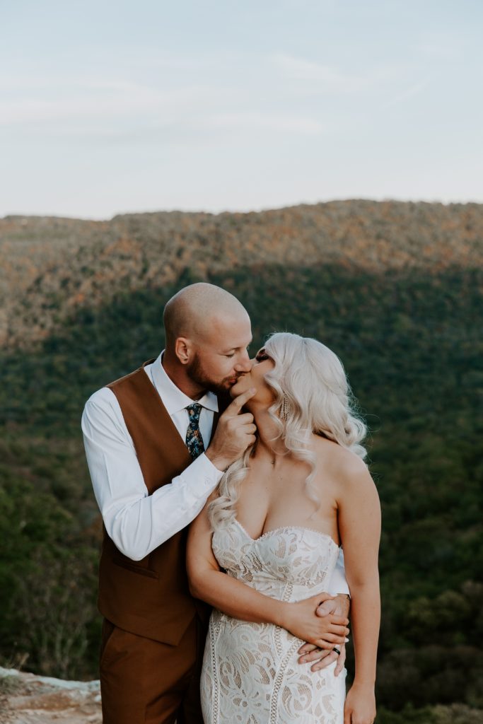 A man gently giving his partner a a kiss while they are holding hands with the Ozarks in the background during their all day elopement