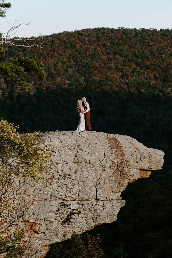 A couple sharing a kiss on their wedding day standing on a cliff as the last light of the day shines on them during their hiking elopement in the Ozarks