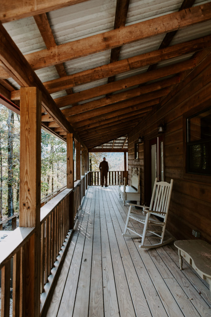 Man standing at the end of a cabin porch waiting for his partner to tap him on his shoulder during their cabin first look in the Ozarks