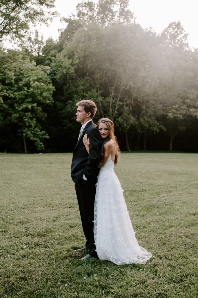 The bride holding on to the back of her partner as he looks off into the distance during their summer Tennessee wedding