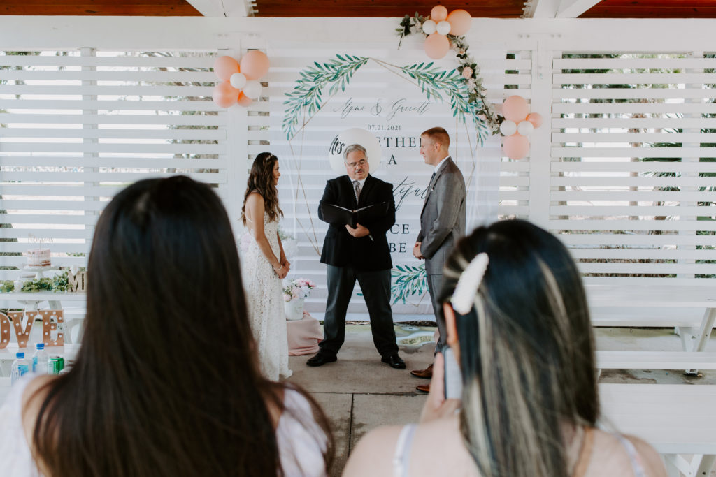 Couple standing with the officiant in front of a sign as the officiant is speaking and their guests are standing around watching