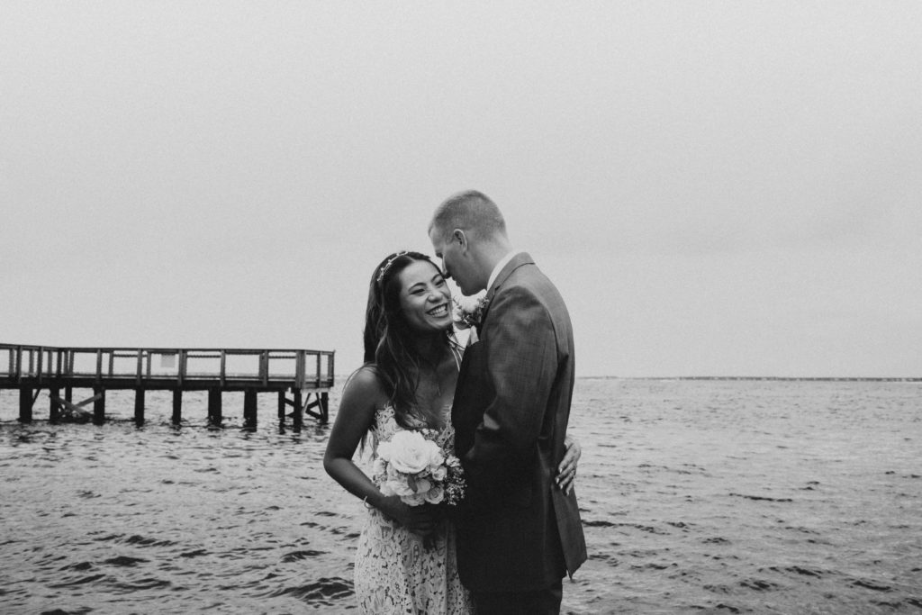 Man whispering into his partners ear as she is laughing and holding on to him while they are standing on a dock