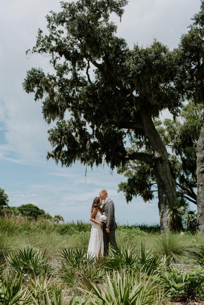 Couple standing on a pathway surrounded by greenery and a large tree behind them as the man is giving his partner a kiss on the forehead during their Florida destination wedding