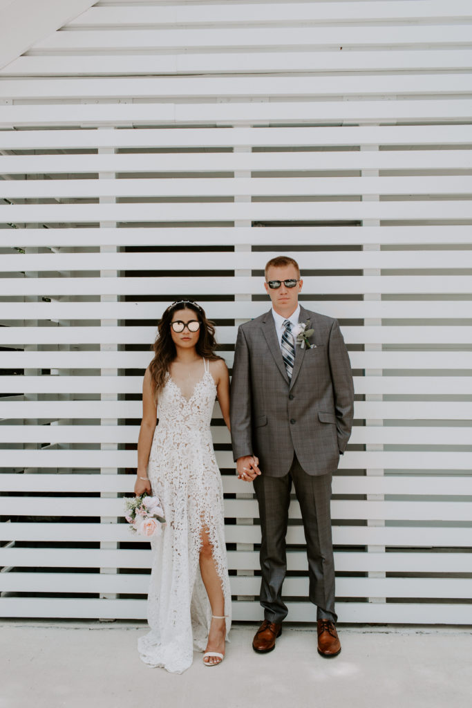 Couple standing side by side holding hands with sunglasses on in their wedding attire looking like the cover of a magazine during their wedding portraits of their Florida elopement