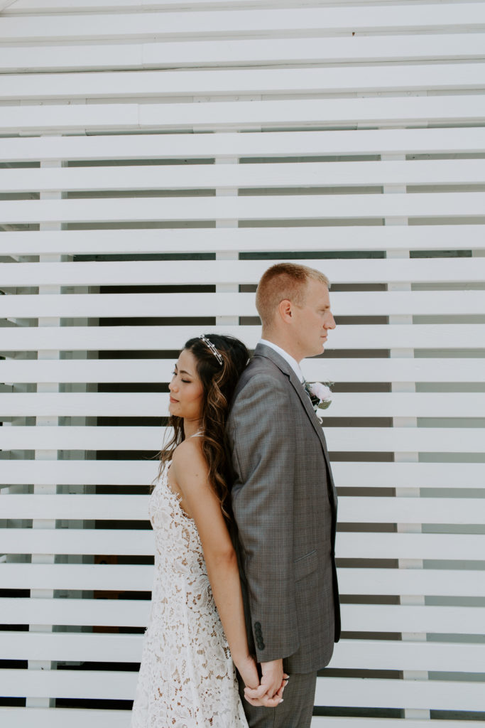 Newly wed couple standing back to back holding hands and looking off in the direction they are standing in front of a white wall during their Destin Florida elopement