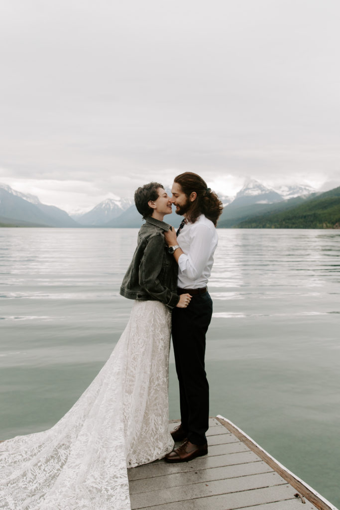 Man bringing his partner in for a kiss by her jacket as they stand on a dock with mountains in the background