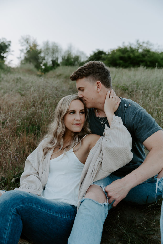 A newly engaged couple sitting in the middle of a field with the woman sitting in her partners lap holding his neck as he gives her a kiss in the head at their seattle engagement photos